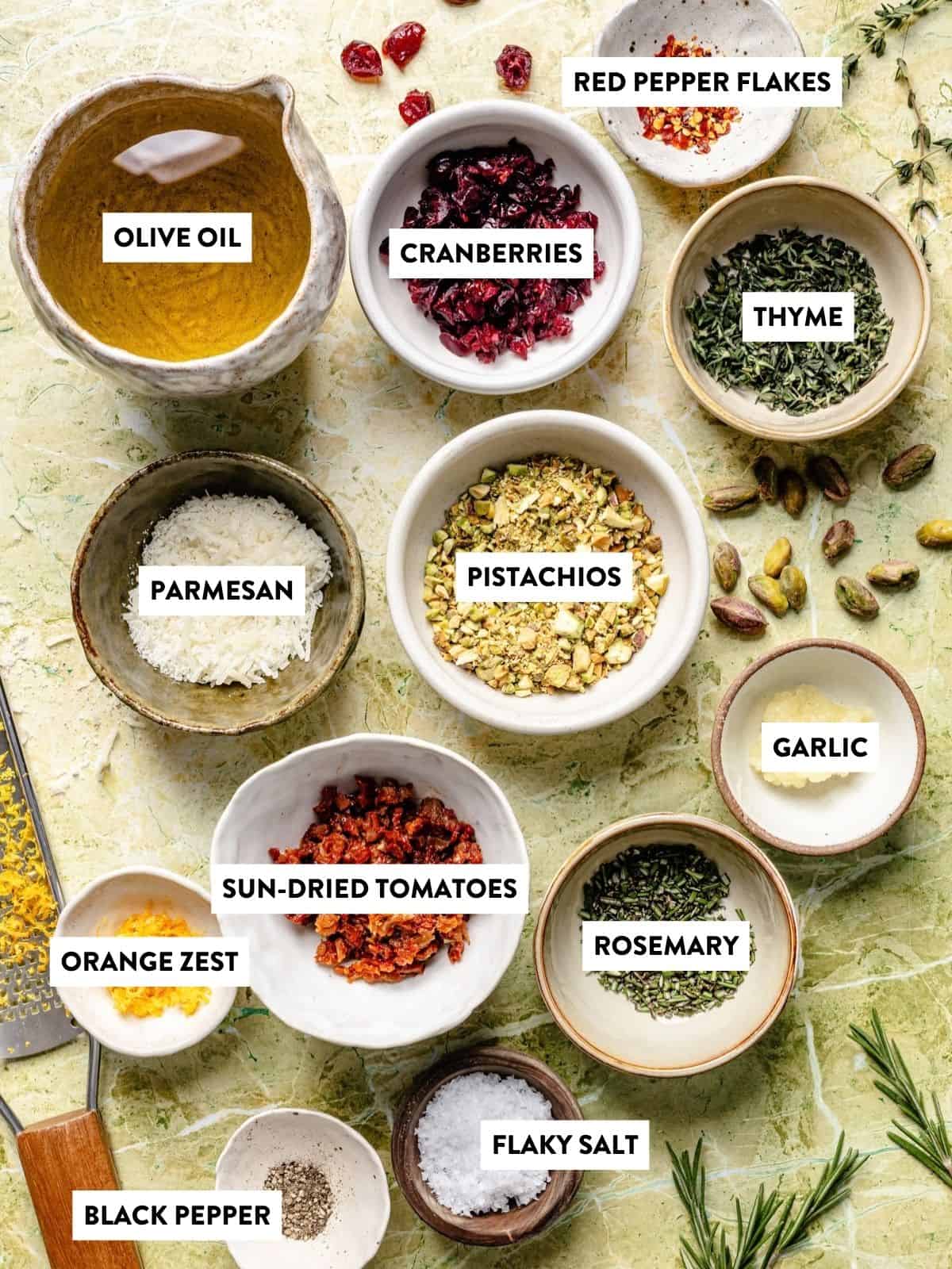 Top-down view of eleven small bowls containing ingredients labeled olive oil, dried cranberries, red pepper flakes, thyme, parmesan, pistachios, garlic, orange zest, sun-dried tomatoes, rosemary, black pepper, and flaky salt for bread dipping oil.